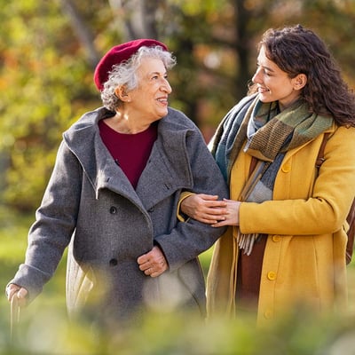A senior grandma smiles as her granddaughter walks arm in arm with her outdoors.