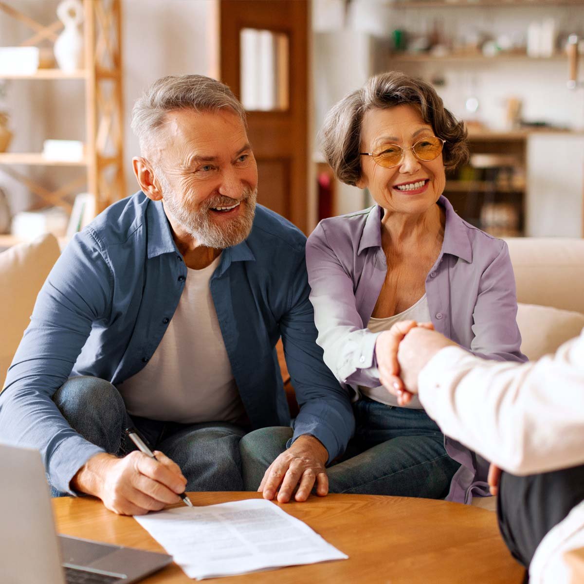 A male senior signs paper work and a female senior shakes the hand of a realtor.