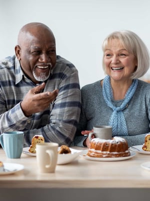 A group of seniors smile and embrace while eating cake and drinking coffee.