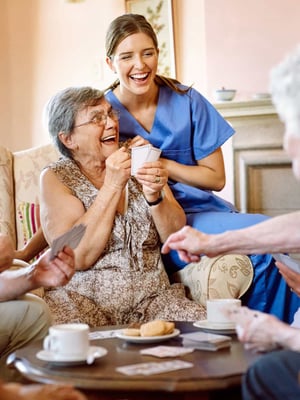 A group of seniors and a nurse laugh and smile while playing a card game.