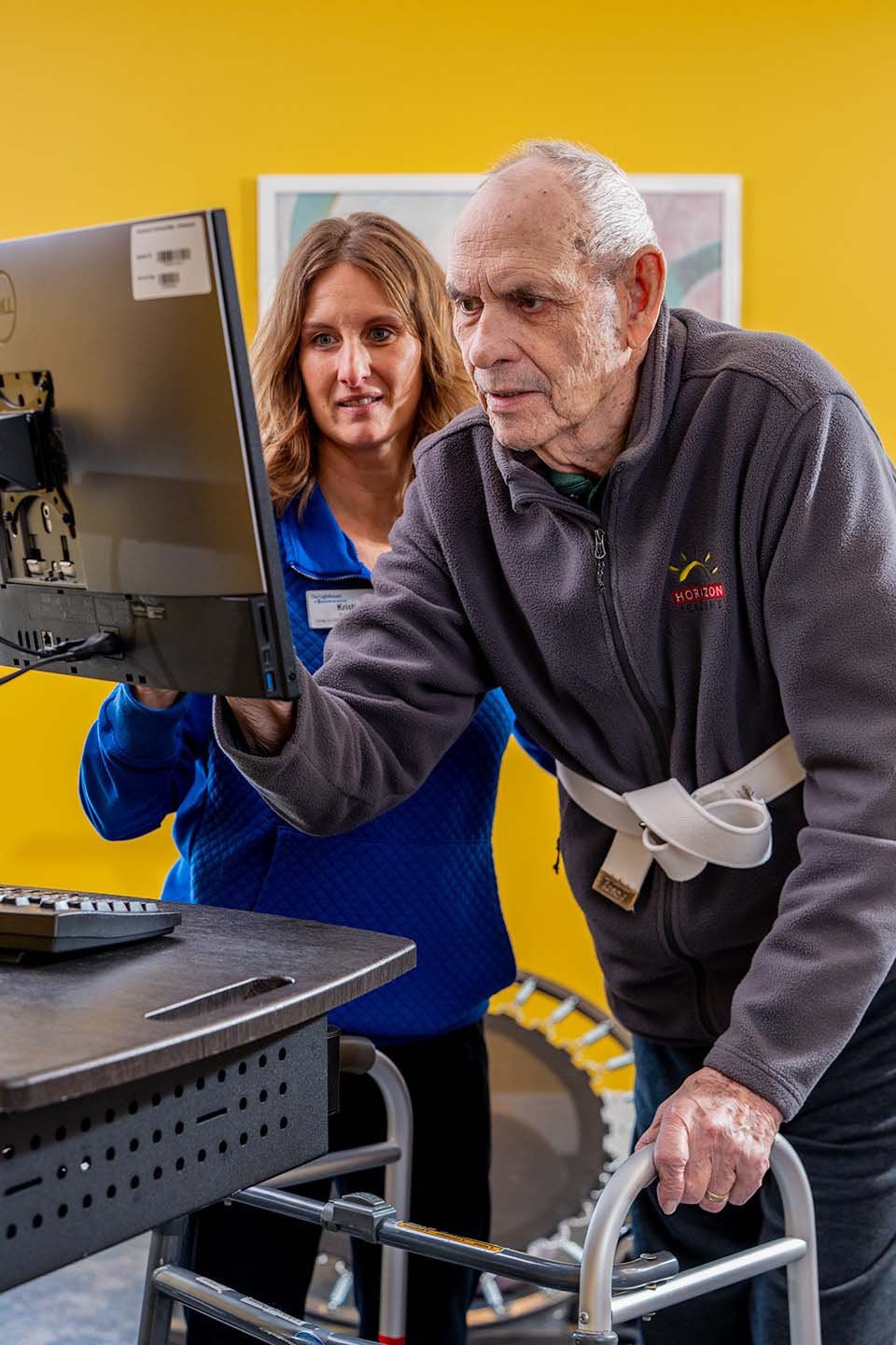 A senior is using a computer with a nurse standing near by.