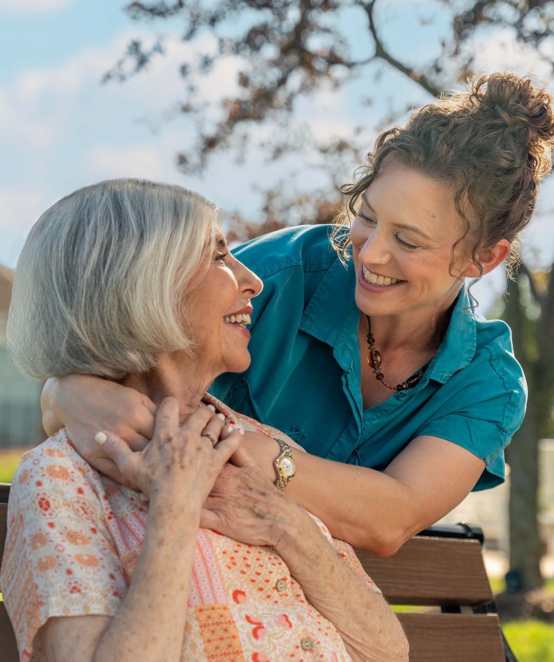 Female senior and adult child sitting on park bench smiling at each other.