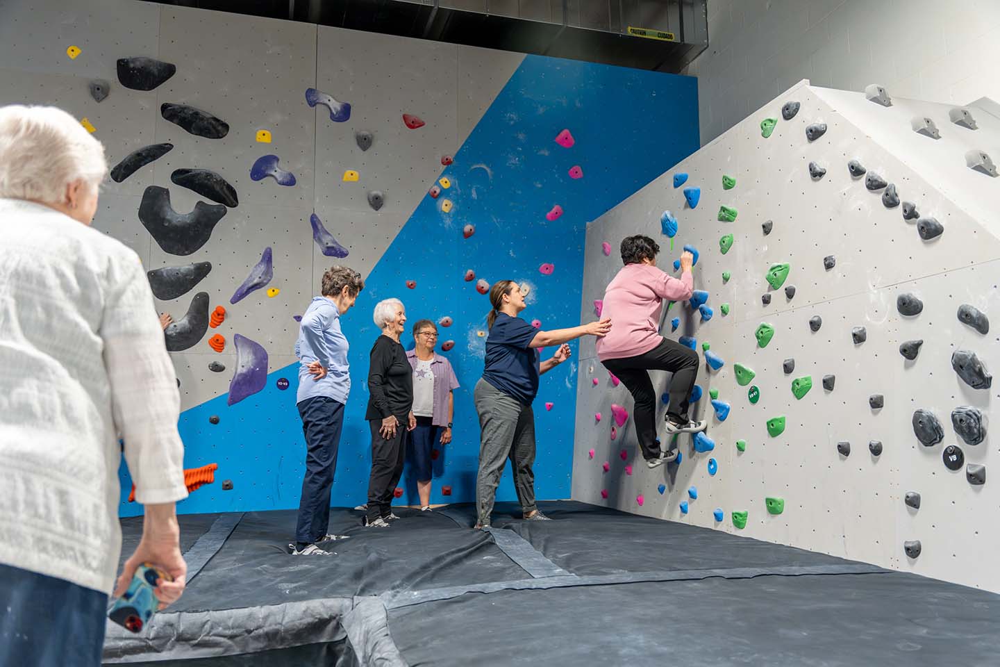 A senior woman climbs an indoor rock wall with the guidance of an instructor.