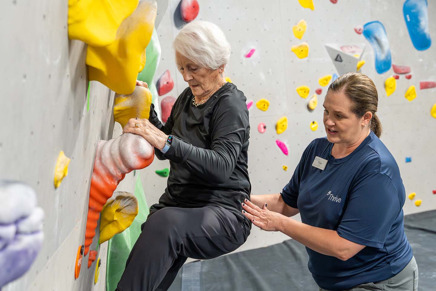 A senior woman climbs an indoor rock wall with the guidance of an instructor.