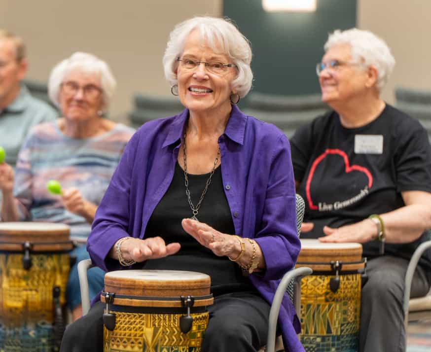 A female senior smiles as she plays a hand drum to music. 